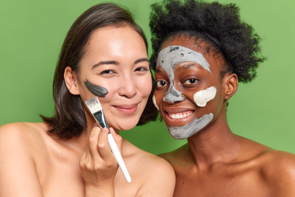 Pleased mixed race women apply beauty masks use cosmetic brush stand closely to each other have clean healthy skin pose bare shoulders against vivid green background. Facial care and cleansing